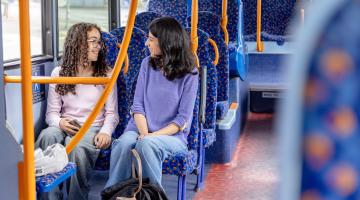A mother and daughter sat on a bus with patterned seats looking towards each other and smiling.