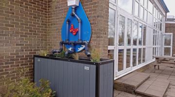 Large rectangular tub with four plants in it sits underneath a drainpipe, which has been split off into three pipes that are dispensing rainwater into the planter. Pipes are in front of a large graphic raindrop with a red butterfly, against a brick wall, which seems to be the back of a school building.