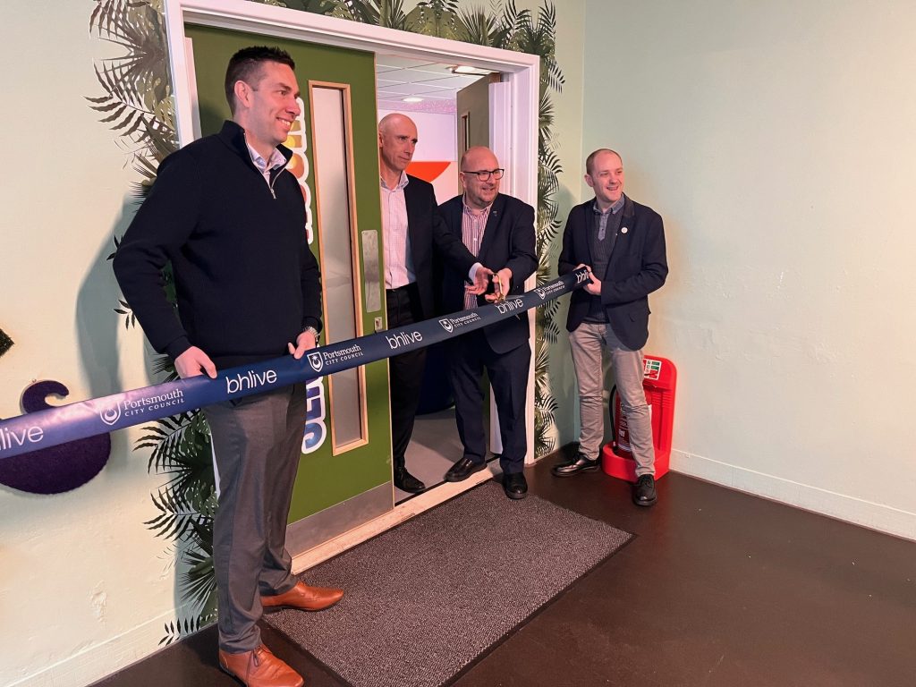 four men holding a ribbon outside a new sensory room, about to cut it and declare the room open